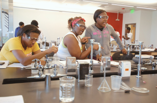 Three students wearing safety goggles work collaboratively at a laboratory table filled with glassware and scientific equipment.